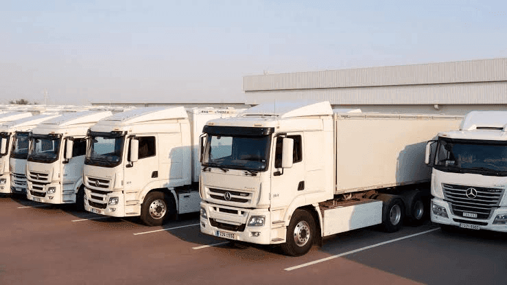 A row of white Mercedes-Benz semi-trailer trucks is parked in a lot near a large industrial building under a clear sky.