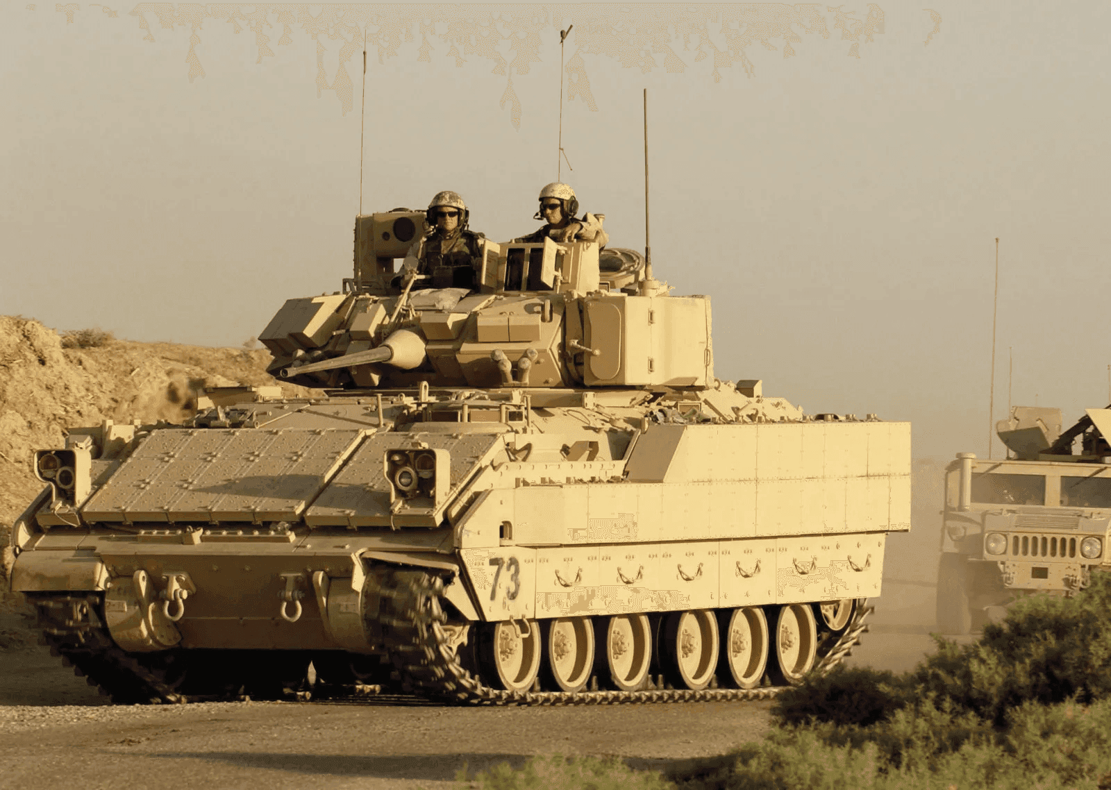 A military armored vehicle (Bradley Fighting Vehicle) with two soldiers riding in the open turret, driving on a desert road with a Humvee following behind.