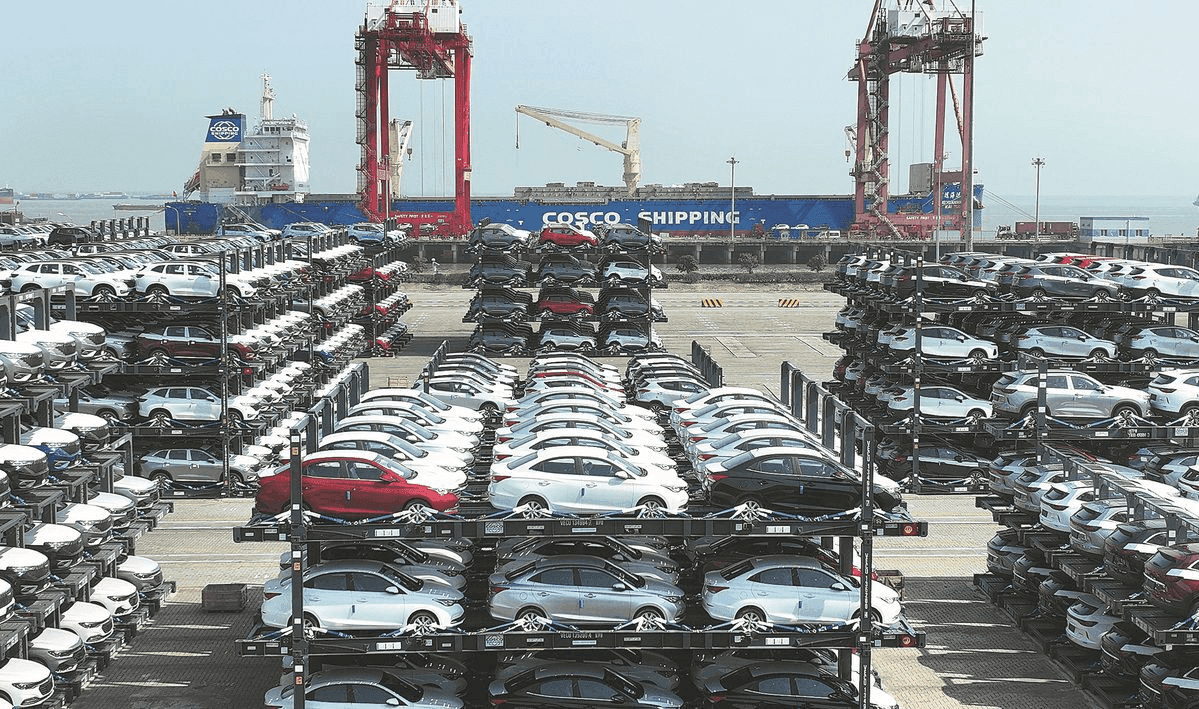 Rows of new cars parked at a port terminal with a COSCO Shipping cargo vessel and red gantry cranes in the background, ready for export.