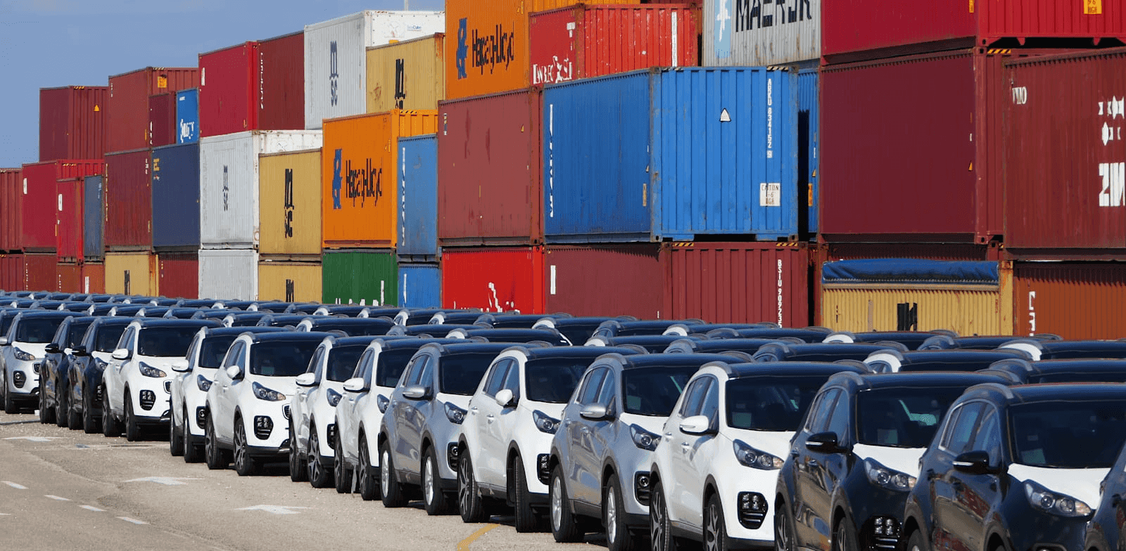 Rows of white and black SUVs parked at a shipping yard with tall stacks of colorful shipping containers in the background.